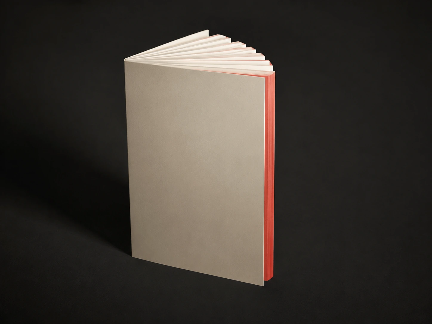 A perfect-bound corporate report standing on its spine, bone cover, cracked open so the text block fans, with a coral sprayed fore-edge