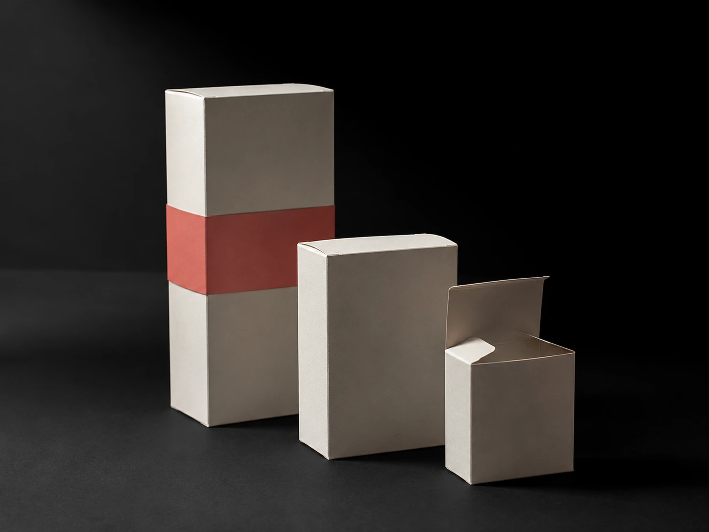 Three folding cartons in uncoated bone board at different scales, one with its tuck flap open, a coral belly band on the tallest