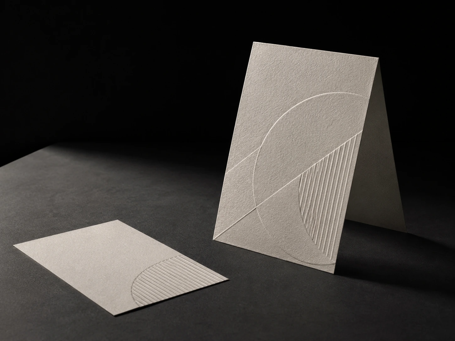 A tent-folded menu card in heavy cotton stock standing on a dark table, deep letterpress relief catching raking light, a second flat card beside it